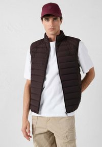 Bruine pufferbodywarmer met een ritssluiting aan de voorkant, horizontaal doorgestikt patroon en een logopatch. Draagt over een wit T-shirt en gecombineerd met kaki broek.