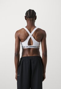 Vit sport-bh med korsat bakdesign och elastiskt band som har texten "UNDER ARMOUR". Matchas med svarta träningsshorts.