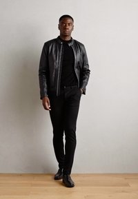Zwarte leren jas met een ritssluiting aan de voorkant, gecombineerd met een zwarte trui en slim-fit zwarte broeken. Model draagt zwarte dresschoenen.