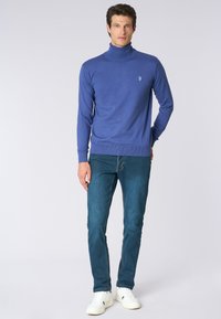 Maglione turtleneck blu realizzato in tessuto liscio, con un piccolo emblema sul petto, abbinato a jeans slim-fit e sneakers bianche.