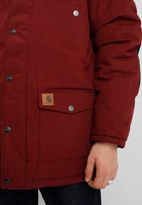 Bordeaux pufferjack met drukknopen, voorzien van een voorzak met een bruine label en zilveren hardware accenten.