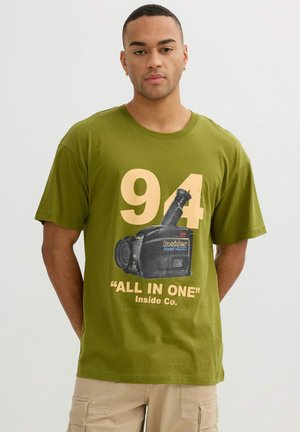 Hombre con camiseta verde oliva con gran "94" y gráfico de cámara de video vintage, pantalones cargo beige, de pie frente a un fondo liso.