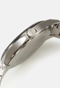 Reloj de acero inoxidable plateado con una caja redonda, corona lateral y superficie texturizada. El diseño presenta un estilo minimalista y acentos sutiles.