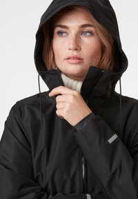 Zwarte waterdichte jas met capuchon, met een hoge kraag, ritssluiting en grijze accenten op de mouwen. Gladde textuur.