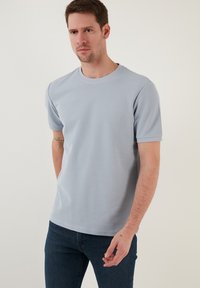 Tricou scurt cu mânecă scurtă, de culoare albastru deschis, cu decolteu rotund, material texturat și croială lejeră, purtat cu blugi de culoare albastru închis.