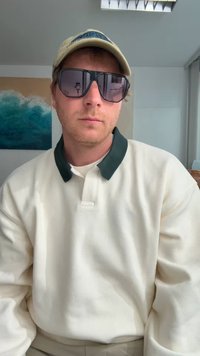 Hvid langærmet polo med en mørkegrøn krave, parret med oversized sorte solbriller og en lysefarvet kasket, indendørs med abstrakt kunst i baggrunden.