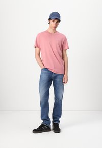 Camiseta rosa de manga corta con cuello redondo, combinada con jeans azul claro desgatados y zapatillas negras. Se lleva una gorra azul en la cabeza.
