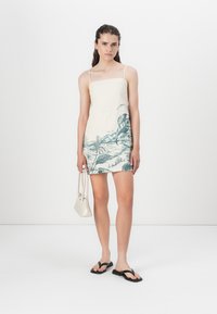 Linnen mini-jurk, crème met groene tropical print. Bevat dunne bandjes, een rechte snit en zakken. Gepaard met zwarte slippers en een witte tas.