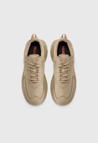 Beige athletic sneakers with a textured fabric upper, lace-up closure, and a thick, rounded sole. Black insole with red logo detail.