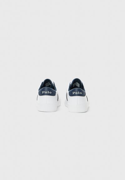 Une paire de baskets blanches avec des talons de couleur bleu marine affichant le texte "Polo", vue de l'arrière sur un fond blanc uni.