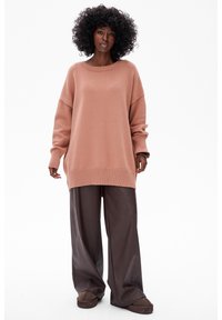 Różowy, oversize'owy sweter dzianinowy z prążkowanym wykończeniem, zestawiony z luźnymi brązowymi skórzanymi spodniami oraz brązowymi butami wsuwanymi. Teksturowany, swobodny fason.
