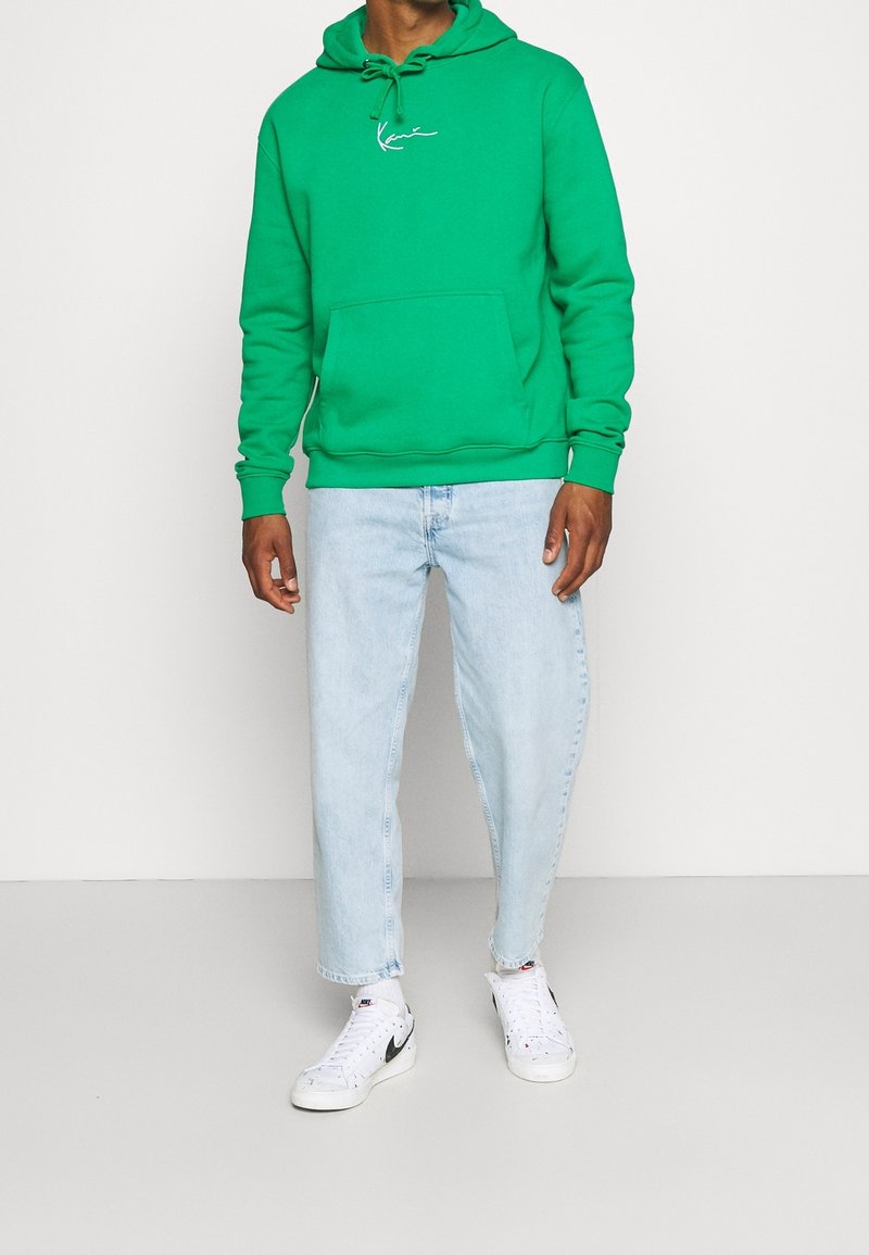 Sudadera verde con un bolsillo frontal y capucha ajustable, combinada con vaqueros rectos de color azul claro y zapatillas blancas.