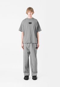 Camiseta gris de gran tamaño con una etiqueta negra en el pecho, combinada con pantalones de chándal grises que presentan un detalle de cadena lateral y un corte relajado. Zapatos negros.