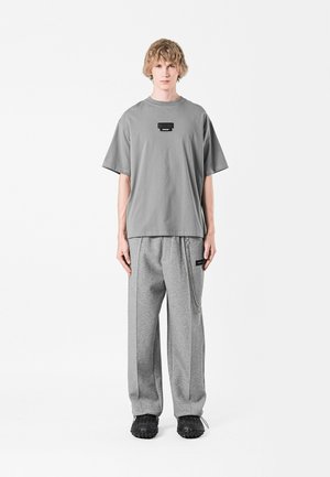 Camiseta gris de gran tamaño con una etiqueta negra en el pecho, combinada con pantalones de chándal grises que presentan un detalle de cadena lateral y un corte relajado. Zapatos negros.