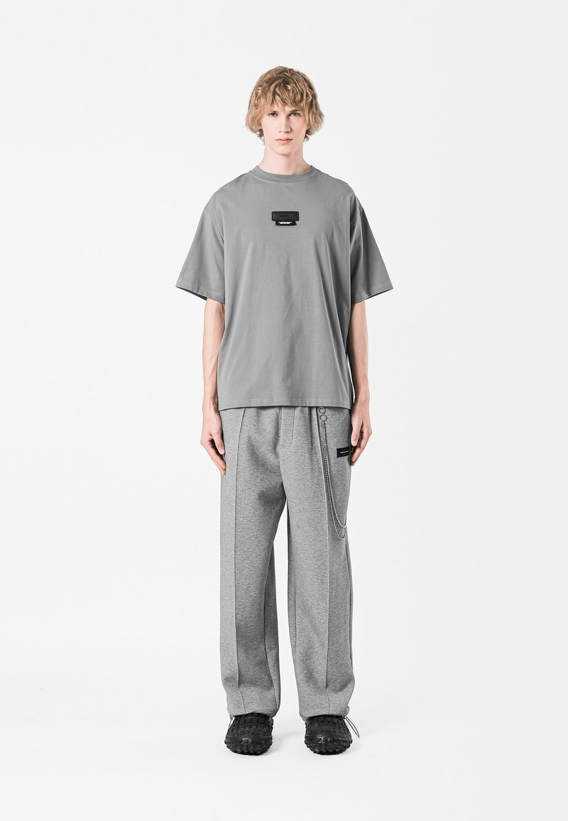 Camiseta gris de gran tamaño con una etiqueta negra en el pecho, combinada con pantalones de chándal grises que presentan un detalle de cadena lateral y un corte relajado. Zapatos negros.