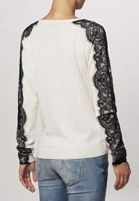 Creamfärgad tröja med svarta spetsdetaljer på axlar och ärmar, ribbade muddar och midja, samt en avslappnad passform.