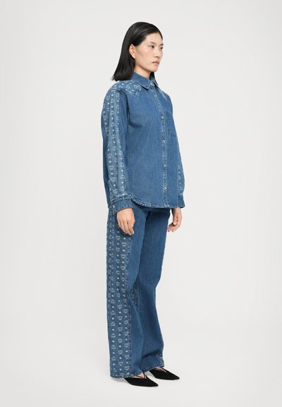 Cămașă din denim cu buzunar, prezentând un model floral embosse pe mâneci; asortată cu blugi cu pantalon larg care afișează detalii similare.