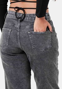 Pantalones de mezclilla gris de cintura alta con un corte relajado, que cuentan con dos bolsillos traseros y un acabado texturizado. La cinturilla tiene un detalle atado en la parte superior.