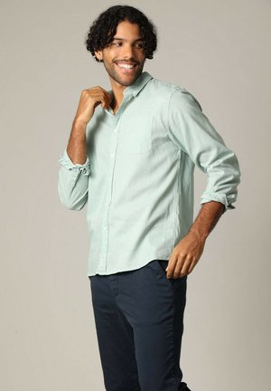 Chemise à manches longues, vert clair, boutonnée, avec une poche poitrine, manches retroussées et coupe décontractée, associée à un pantalon bleu foncé.