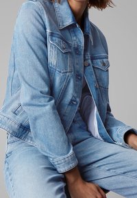 Světle modrá džínsová bunda s knoflíkovým zapínáním, náprsními kapsami a mírně zkráceným střihem. Textura je hladká s jemnými detaily vyblednutí.