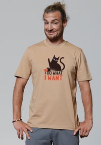 Lichtbeige t-shirt met korte mouwen met een grafiek van een zwarte kat en de tekst "IK DOE WAT IK WIL" in vet rood lettertype. Gladde textuur.