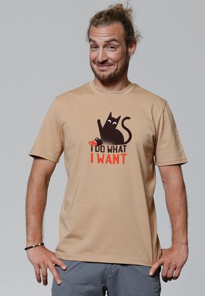 Hellbeige T-Shirt mit kurzen Ärmeln, das ein schwarzes Katzenmotiv und den Text "ICH MACH, WAS ICH WILL" in fetten roten Buchstaben zeigt. Glatte Textur.