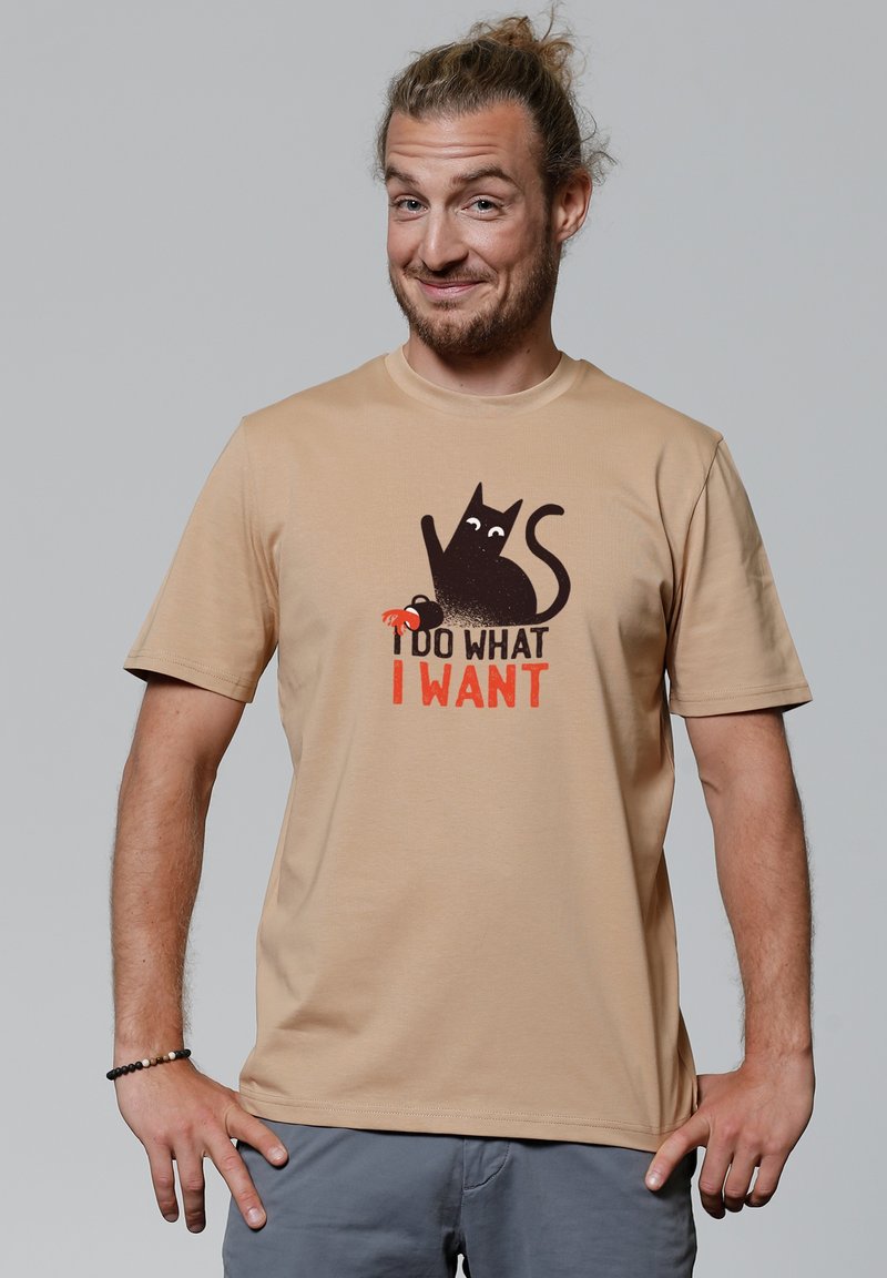 Lichtbeige t-shirt met korte mouwen met een grafiek van een zwarte kat en de tekst "IK DOE WAT IK WIL" in vet rood lettertype. Gladde textuur.