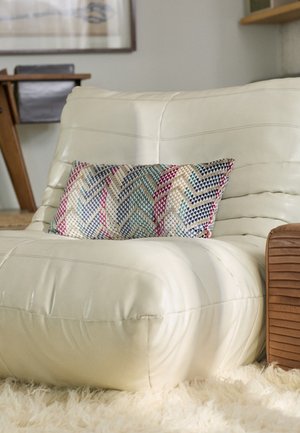 Cream leather armchair with a textured, chevron-patterned pillow in blue, pink, and beige. Wood accent on the side. Soft, shaggy rug beneath.