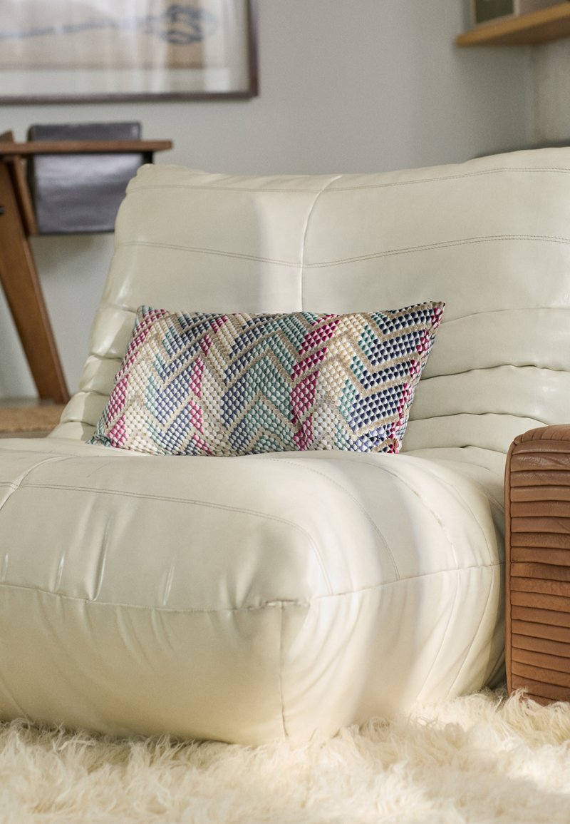 Cream leather armchair with a textured, chevron-patterned pillow in blue, pink, and beige. Wood accent on the side. Soft, shaggy rug beneath.