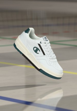 Pantof sport alb și turcoaz cu logo Champion plutind deasupra podelei unei săli de sport cu linii de delimitare colorate.