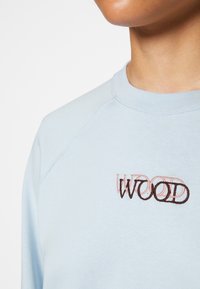 Sudadera azul claro con cuello redondo y mangas ranglán. Presenta el bordado "WOOD" en marrón y rosa en el pecho. Textura suave.