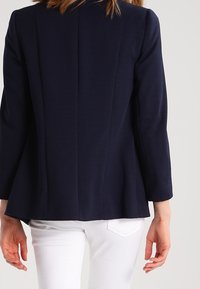 Mörkblå texturerad blazer med långa ärmar och skräddarsydd passform, med en söm bak och subtil peplumdesign. Bärs över vita byxor.