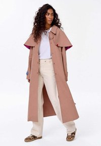 Kobieta z długimi kręconymi włosami, ubrana w beżowy trencz over białą koszulę, kremowe spodnie i trampki w panterkę, stojąca na białym tle.