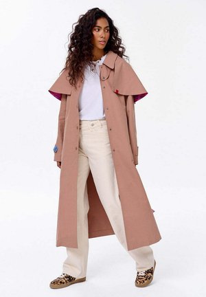 Žena s dlouhými kadeřavými vlasy, oblečená v béžovém trenčkotě přes bílou košili, krémových kalhotách a teniskách s leopardím potiskem, stojící na bílém pozadí.