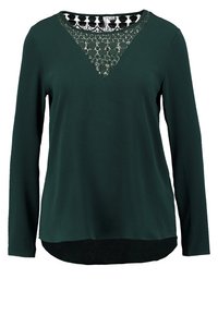 Chemise à manches longues vert foncé avec un détail en dentelle au niveau de l'encolure, un ourlet noir en bas, et une coupe décontractée. Texture de tissu douce.