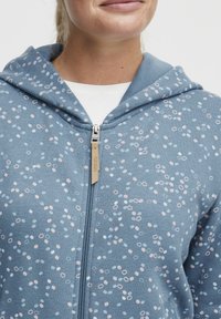 Ljusblå hoodie med dragkedja, prydd med ett spritt mönster av rosa och vita prickiga cirklar. Tyget är mjukt, med en avslappnad passform och huva.
