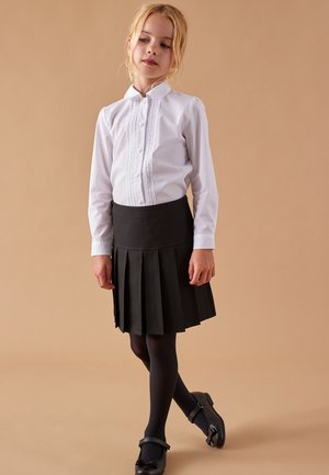 Weißes Langarmshirt mit Knopfleiste und Spitze, kombiniert mit einem schwarzen Faltenrock sowie schwarzen Strumpfhosen und Schuhen.