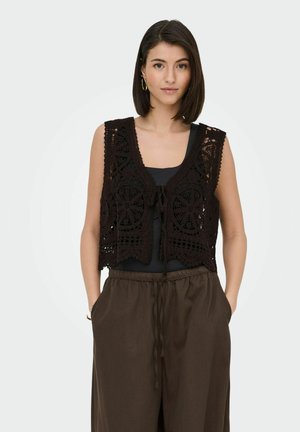 Donna in piedi di fronte, indossa un gilet nero all’uncinetto sopra una maglietta nera e pantaloni marroni larghi con coulisse, con le mani nelle tasche.