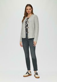 Cardigan en tricot gris clair avec des boutons en bois, associé à un t-shirt graphique et un jean foncé. Les baskets présentent un design beige et noir.