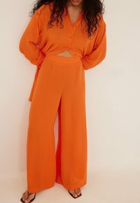 Ensemble en deux pièces orange composé d'une chemise en tissu fluide à manches longues et à l'avant noué, accompagnée d'un pantalon évasé. Tissu lisse avec une coupe décontractée.