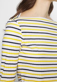 Randig topp med horisontella linjer i vitt, marinblått och gult. Ribbad textur med vid nederkant och åtsittande design.