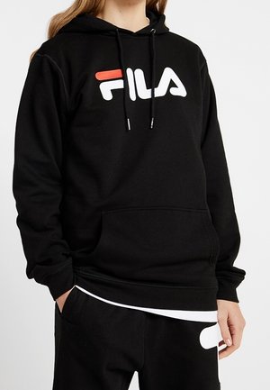 Person iført en sort FILA-hoodie med rødt og hvidt logo, hænderne i lommerne, kombineret med sorte bukser med hvidt design.