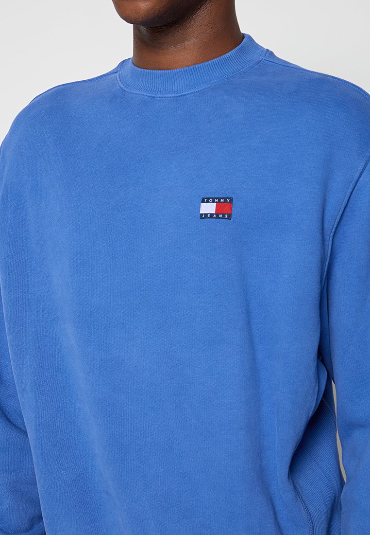 Niebieska bluza z bawełny, z okrągłym dekoltem, elastycznymi mankietami i małą naszywką z logo w kolorach czerwonym, białym i niebieskim na piersi.