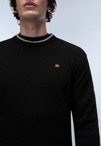 Czarny sweter z dzianiny z okrągłym dekoltem, z szarym paskiem na kołnierzyku oraz małym czerwono-białym emblematem na piersi.