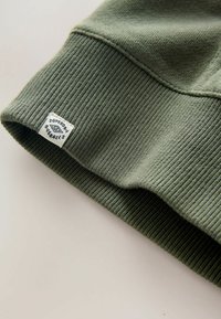 Polsino in maglia a coste verde oliva, con un'etichetta tessuta che mostra "Superdry Overalls" in blu su sfondo bianco.