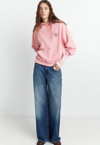 Felpa rosa con tasca frontale e logo a forma di cuore; abbinata a jeans larghi blu e scarpe marroni. Texture lisce e vestibilità casual.