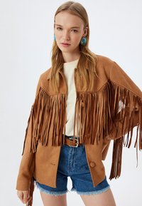 Bruine suède jas met lange franjes, knoopsluiting en een kraag, gecombineerd met een beige top en denim shorts. Blauwe oorbellen zichtbaar.