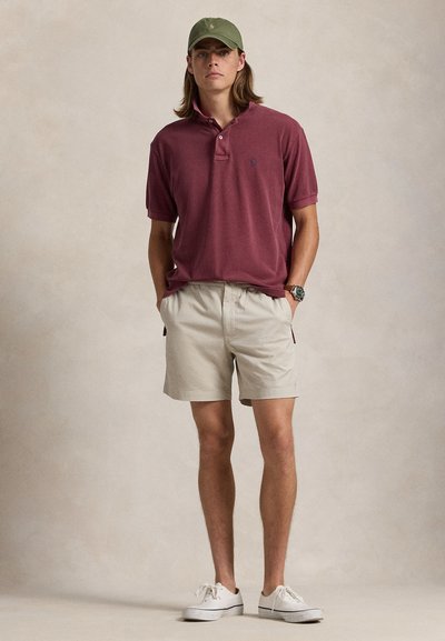 Vinröd pikétröja med korta ärmar och krage; beige shorts; vita sneakers; grön keps; diskret logotypdetalj på tröjan.