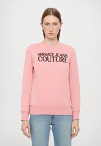 Rozā džemperis ar melnu "VERSACE JEANS COUTURE" logotipu, apaļā kaklā, gariem piedurknēm, ribotām aprocēm un apakšmalu, izgatavots no mīksta auduma.