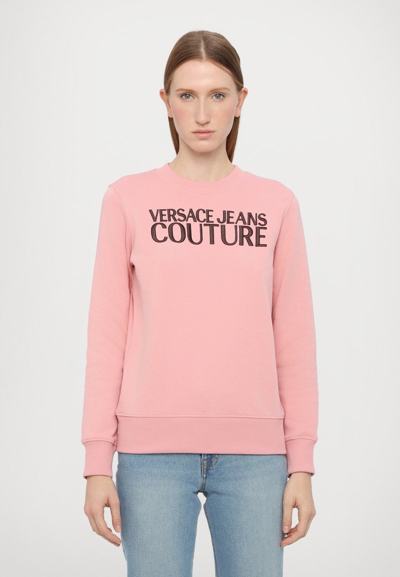 Rozā džemperis ar melnu "VERSACE JEANS COUTURE" logotipu, apaļā kaklā, gariem piedurknēm, ribotām aprocēm un apakšmalu, izgatavots no mīksta auduma.
